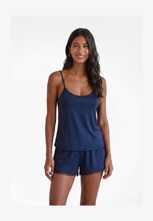 Frau lächelt, trägt ein dunkelblaues Spaghettiträger-Top und passende Shorts mit transparentem Besatz, steht vor einem schlichten weißen Hintergrund.