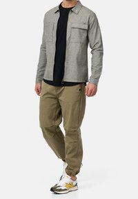 Homme portant une veste grise à fermeture éclair par-dessus un t-shirt noir, un pantalon cargo kaki et des baskets blanches avec des touches jaunes, debout avec un pied en avant.