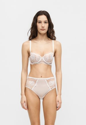 Femme debout portant un soutien-gorge en dentelle blanche assorti et une culotte taille haute, expression neutre, fond gris clair uni.