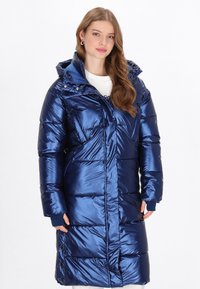 Hosszú kék puffertakaró, fényes anyagból készült, kapucnival, cipzárral elöl és bordás mandzsettákkal. Kiemelkedő a steppelt, párnázott kialakítása.