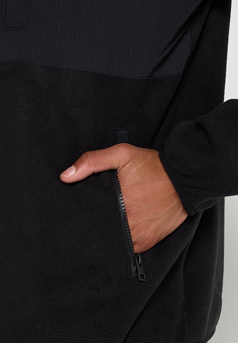 Pull-over en polaire noire avec une partie supérieure côtelée, comportant une poche zippée à l'avant et une surface douce et texturée.