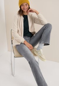 Lächelnde Frau in einer senfgelben Mütze, cremefarbener Fleecejacke, dunklem Shirt, grauen Jeans und beigen Sneakers sitzt lässig auf einem beigen Stuhl vor einem schlichten Hintergrund.