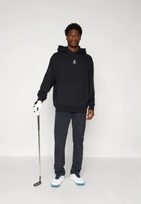 Μαύρο φούτερ με κουκούλα, μπροστινή τσέπη και λογότυπο, σε συνδυασμό με παντελόνι σκούρου γκρι. Το μοντέλο κρατάει μπαστούνι γκολφ και φοράει λευκά παπούτσια γκολφ με μπλε λεπτομέρειες.