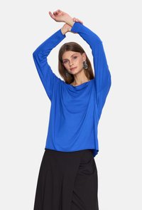 Niebieska bluzka z długim rękawem i luźnym fasonem, wykonana z miękkiego materiału; połączona z czarną spódnicą.