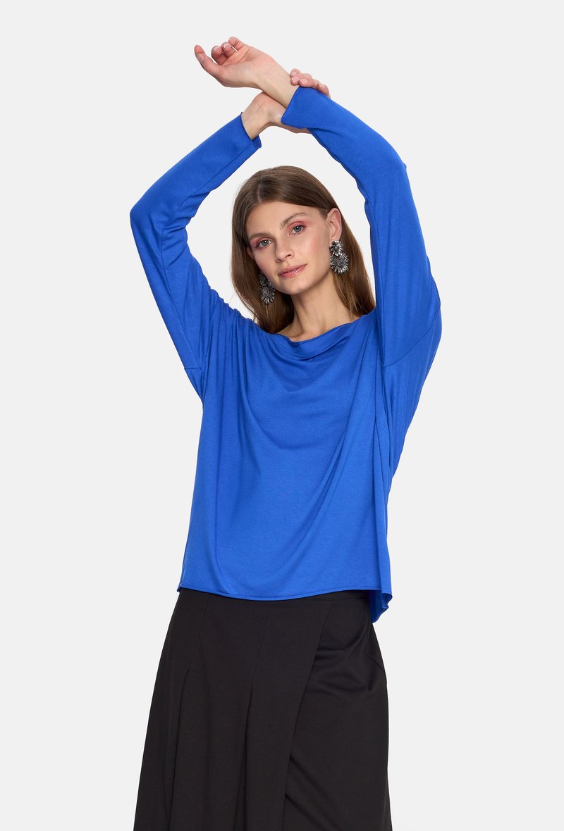 Niebieska bluzka z długim rękawem i luźnym fasonem, wykonana z miękkiego materiału; połączona z czarną spódnicą.