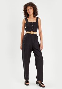 Black jumpsuit with square neckline, button detail, and side pockets. Woven beige belt with round buckle accents the waist. Flat black sandals.