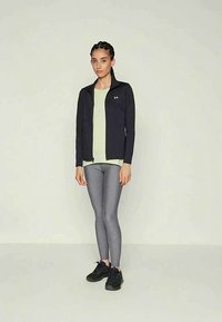 Nő áll, fekete cipzáras dzsekiben, világos zöld pólóban, szürke leggingsben és fekete sportcipőben, egy sima világos háttér előtt.