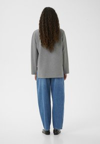 Maglia grigia a maniche lunghe con taglio dritto, abbinata a pantaloni in denim blu larghi. Capelli ricci che scendono fino a metà schiena. Scarpe nere visibili.