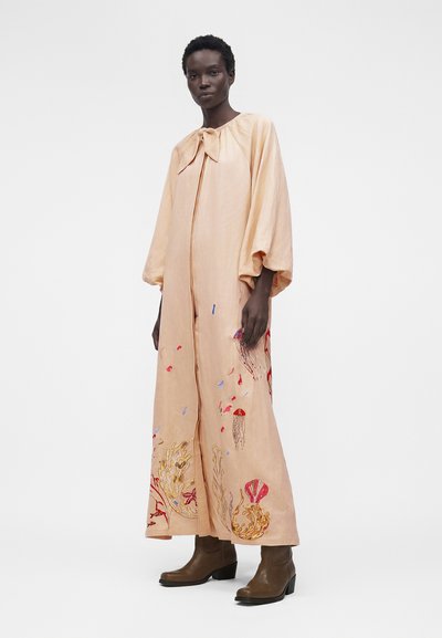 Γυναίκα που φοράει ένα μακρύ ροδακινί φόρεμα με μπαλούν μανίκια και πολύχρωμη κεντητή απεικόνιση θαλάσσιας ζωής, συνδυασμένο με καφέ μποτάκια μέχρι τον αστράγαλο.