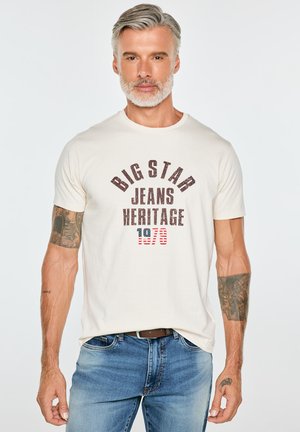 Mężczyzna w średnim wieku z siwymi włosami i brodą, ubrany w białą koszulkę "Big Star Jeans Heritage 1970" i niebieskie dżinsy, pokazujący tatuaże na obu ramionach.