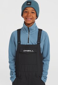 Schwarze Latzhosen von O'Neill mit verstellbaren Trägern, kombiniert mit einem hellblauen Fleece-Zip-Up-Oberteil und einer passenden blauen Strickmütze.