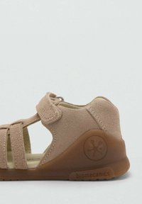 Beige suède sandaal met klittenband, open zijkanten en rubberen zool. Kenmerkt zich door branding op de zool en textuuraccenten van suède.