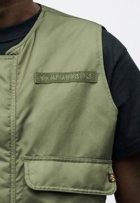 Κοντινή λήψη τεχνικού γιλέκου σε ελιά απόχρωση με τσέπη με καπάκι και το έμβλημα "Alpha Industries", φορεμένο πάνω από μαύρο πουκάμισο.
