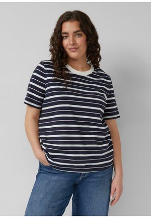 Frau mit lockigem Haar, die ein marineblau-weiß gestreiftes kurzärmeliges Shirt und blaue Jeans trägt, mit einer Hand in der Tasche stehend vor einer grauen Wand.