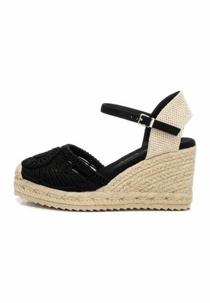 Sandalia de cuña trenzada negra con upper texturizado, correa ajustable en el tobillo y plataforma envuelta en yute. Patrón circular distintivo en la zona de los dedos.