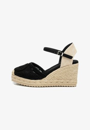 Sandalia de cuña trenzada negra con upper texturizado, correa ajustable en el tobillo y plataforma envuelta en yute. Patrón circular distintivo en la zona de los dedos.
