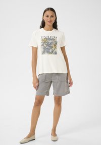 T-shirt crème avec imprimé floral, texte "LES FLEURS", assorti à un short à rayures verticales noir et blanc. Tissu lisse, coupe décontractée.
