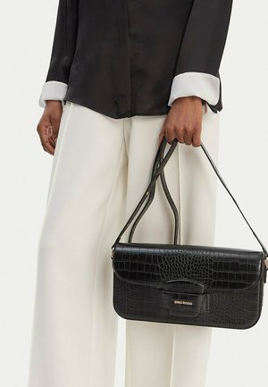 Osoba držící černou kabelku s krokodýlím vzorem a dvojitými pásky, oblečená v černém topu s bílými manžetami a bílých širokých kalhotách.