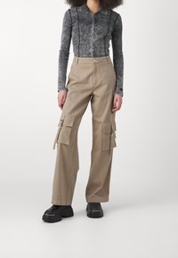 Béžové cargo kalhoty s několika velkými kapsami, vyrobené z odolné tkaniny, v kombinaci se šedou košilí s dlouhým rukávem a kontrastním prošíváním.