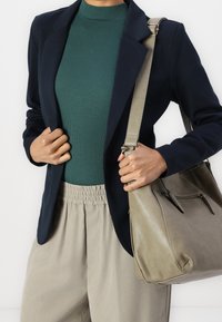 Blazer bleu marine porté sur un haut côtelé vert, associé à un pantalon beige à taille élastique. Un sac gris clair à fermeture éclair est tenu dans une main.
