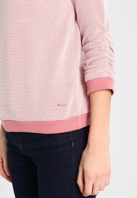 Suéter texturizado de un suave color rosa con un patrón de panal, puños ajustados y un dobladillo acanalado. Acento del logo dorado cerca de la parte inferior izquierda.