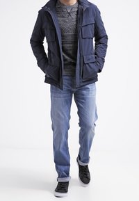 Veste zippée bleu marine avec plusieurs poches, portée sur un pull gris. Un jean bleu clair et des baskets noires complètent la tenue.