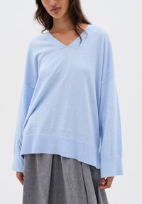 Frau trägt einen hellblauen V-Ausschnitt Oversized-Pullover mit weiten Ärmeln und einen grauen plissierten Rock vor einem einfarbigen Hintergrund.