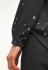 Chemise noire avec un motif de petits oiseaux blancs, munie d'un poignet boutonné et d'une texture de tissu douce. Col visible et couture détaillée.