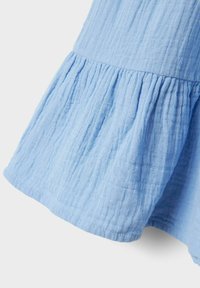 Tissu bleu clair, froissé, avec un ourlet évasé et une taille froncée. La texture est douce et légère. Aucun motif ou matériel visible.