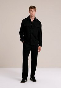 Svart skjorta med knappar och två bröstfickor, kombinerad med matchande byxor. Modellen bär svarta sneakers med vita ränder. Enkel och minimal design.