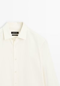 Camicia a maniche lunghe color crema realizzata in tessuto morbido. Presenta un colletto classico, chiusura con bottoni e una leggera lucentezza, con un'etichetta visibile all'collo.