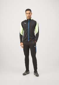 Conjunto deportivo negro de manga larga con acentos en verde lima y azul, frente con cremallera y pantalones ajustados con franjas laterales.