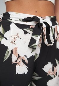 Svarte blomstrete bukser med knytebelte i midjen, store hvite og rosa blomster, grønne blader og et teksturert stoff.