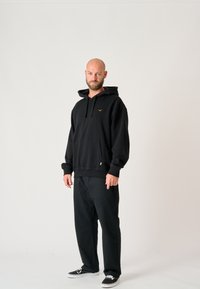 Black hooded sweatshirt with a front pocket and gold logo, paired with loose-fitting black pants and black shoes. Smooth texture, casual design.