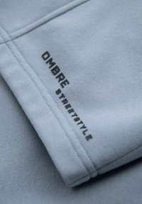 Un textile bleu clair avec une texture douce, présentant un texte imprimé en noir "OMBRE STREETSTYLE" le long du bord. Détails de couture visibles.