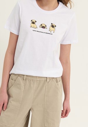 Hvid T-shirt med tre tegneserie-pugs og tekst "Love wrapped in wrinkles" båret med beige bukser på en person mod en ensfarvet baggrund.