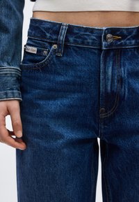 Κοντινό πλάνο ατόμου που φοράει σκούρο μπλε τζιν με ορατό κουμπί, θηλιές για ζώνη και ετικέτα Calvin Klein, σε συνδυασμό με ανοιχτόχρωμο κοντό τοπ.