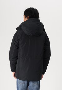 Sort pufferjakke med hætte, der har en glat tekstur, elastiske manchetter og en lige kant, båret over blå jeans.