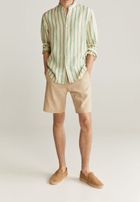 Leichtes Leinenhemd mit grünen Streifen und Mandarin-Kragen, kombiniert mit beigen Shorts und braunen Slippern.