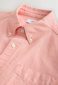 Chemise rose à boutons avec col boutonné et boutons blancs visibles, montrant la poche avant et la texture du tissu.
