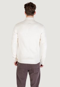 Hombre de espaldas vistiendo un suéter blanco de cuello alto y pantalones marrones frente a un fondo claro y simple.