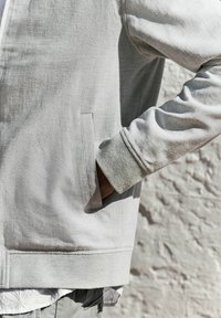 Chaqueta gris claro con tejido texturizado; presenta bolsillos laterales y puños acanalados; estilizada con una camisa en capas por debajo.