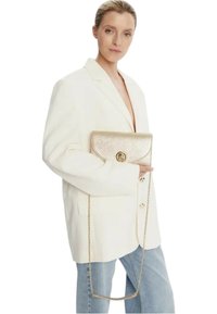 Gouden metallic clutchtas met een overslagklep, kettingriem, gladde textuur, vastgehouden door een persoon die een witte blazer en lichtblauwe jeans draagt.