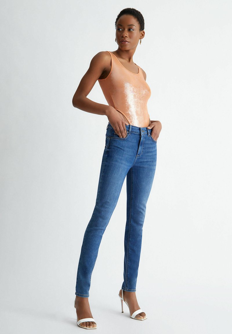 Top à fines bretelles rose doré brillant associé à un jean skinny bleu. Le mannequin porte des sandales à talons blancs, mettant en valeur une silhouette ajustée. Textures et couleurs lisses.