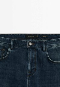 Tummansiniset farkut, joissa on kapeneva malli, edessä taskut, metallinen napitus ja ompeluaksentit. Merkki näkyvissä vyötäröllä.