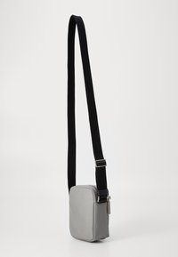 Borsa a tracolla in tessuto grigio con forma rettangolare, design piatto, tracolla nera regolabile e dettagli in metallo argentato sulla cerniera.