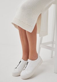 Baskets blanches avec une finition en cuir lisse, fermeture éclair sur le devant et une épaisse semelle blanche, associées à une robe blanche texturée arrivant au genou.