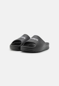 Chanclas negras de goma, con diseño de punta abierta, suela texturizada y el logo de 'Lacoste' en blanco en la parte superior.