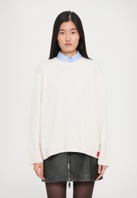 Giovane donna con lunghi capelli neri che indossa un maglione bianco oversize sopra una camicia colletto blu chiaro e una gonna corta in pelle nera con zip.
