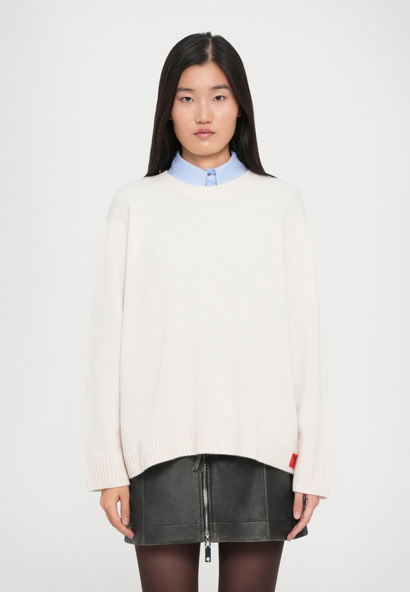 Giovane donna con lunghi capelli neri che indossa un maglione bianco oversize sopra una camicia colletto blu chiaro e una gonna corta in pelle nera con zip.